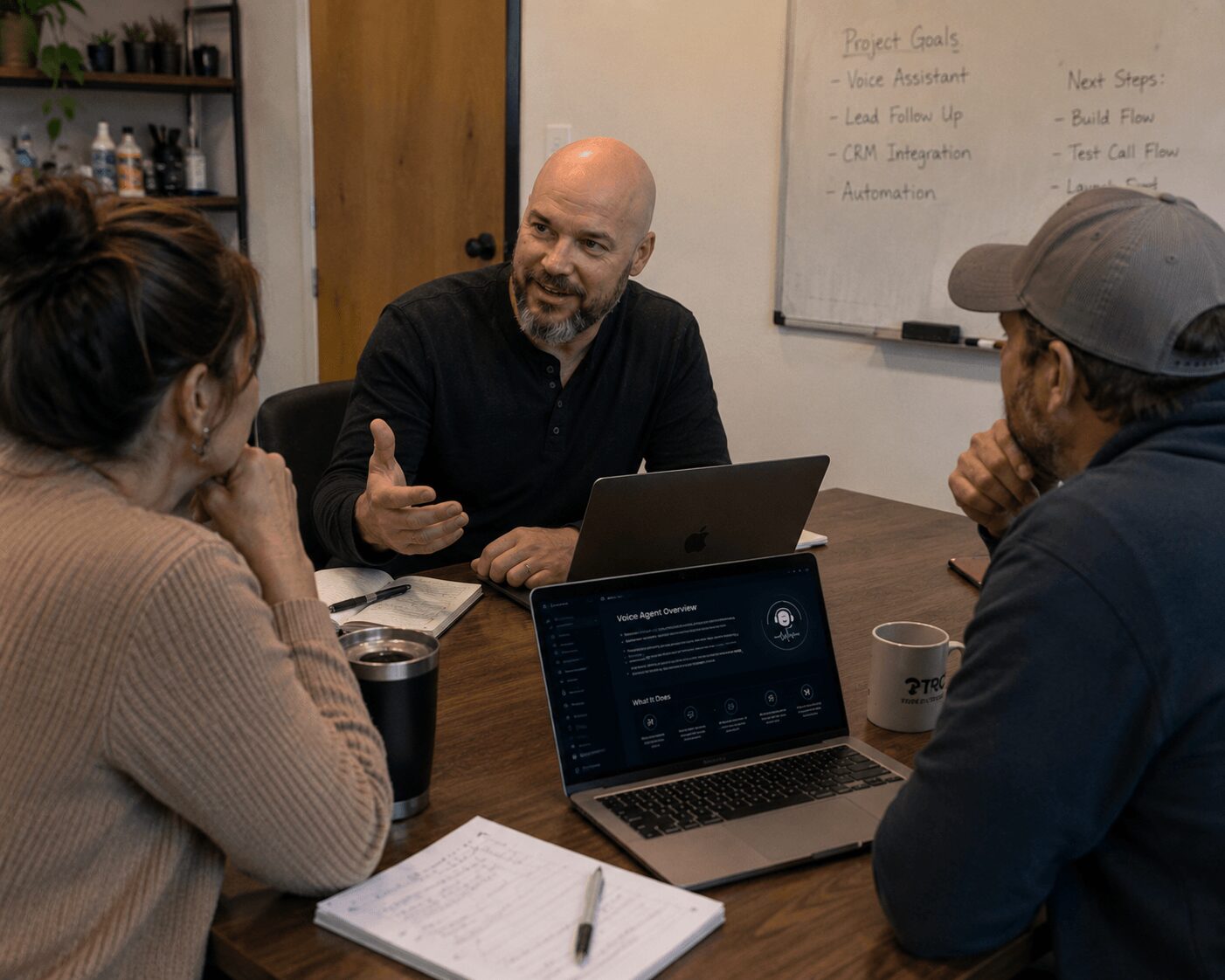 Three people sit around a table in a meeting room, engaged in conversation with laptops and notebooks.