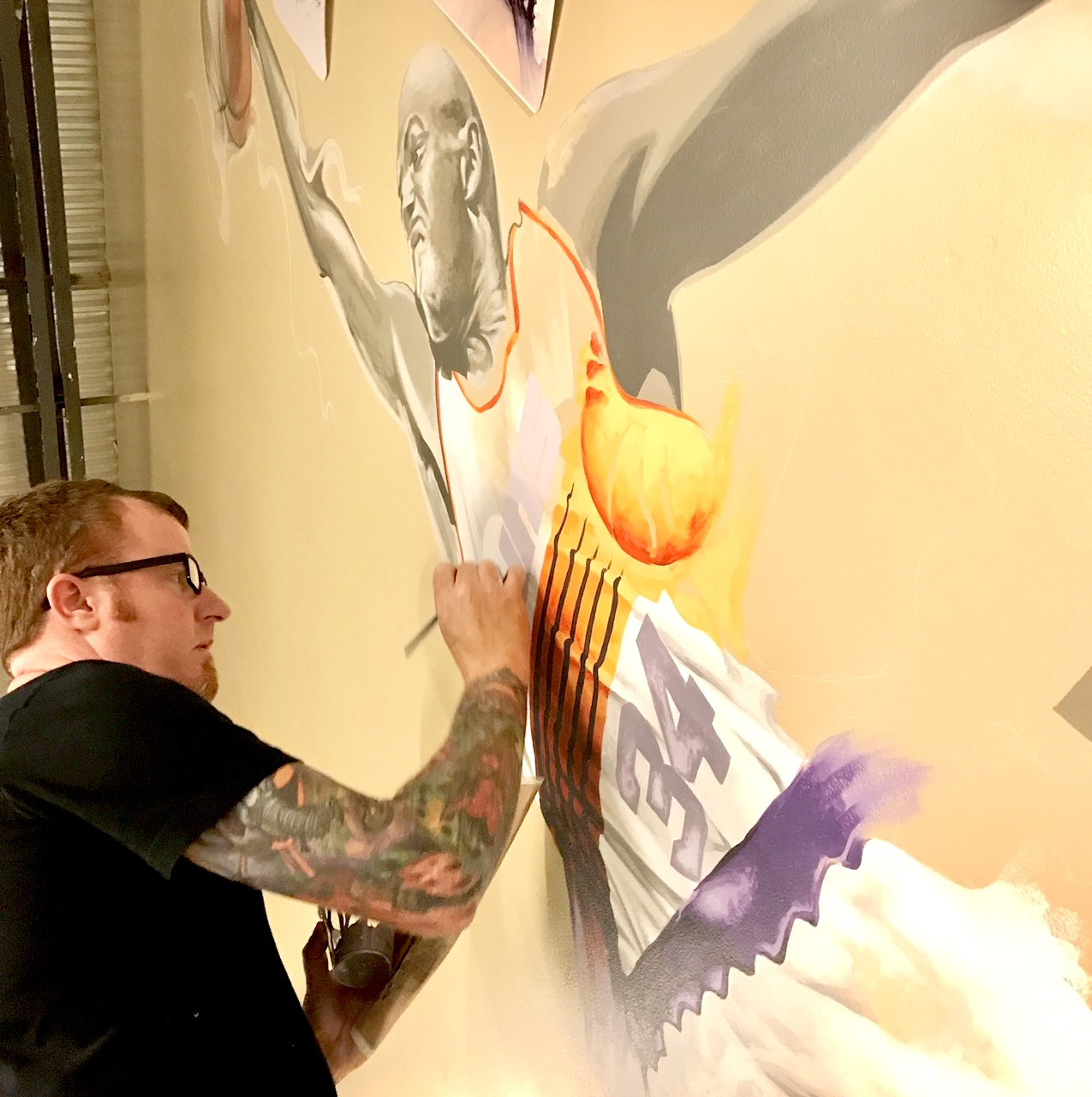 Tattooed man with glasses painting a large mural of a basketball player on a wall.
