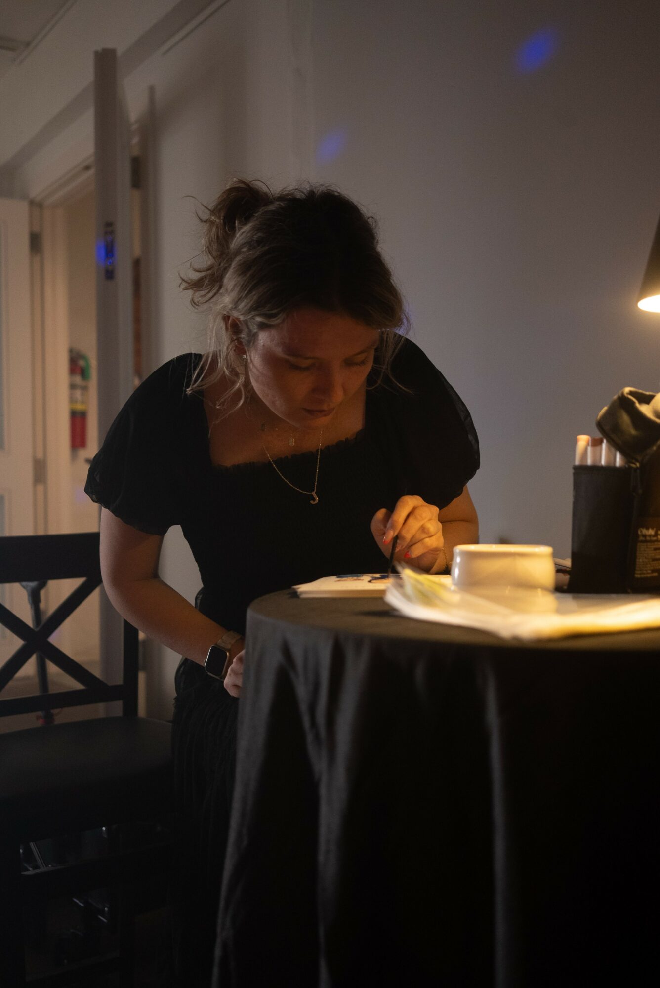 Woman leaning over a table, writing or drawing, with a lamp and objects on the table, in a dimly lit room.