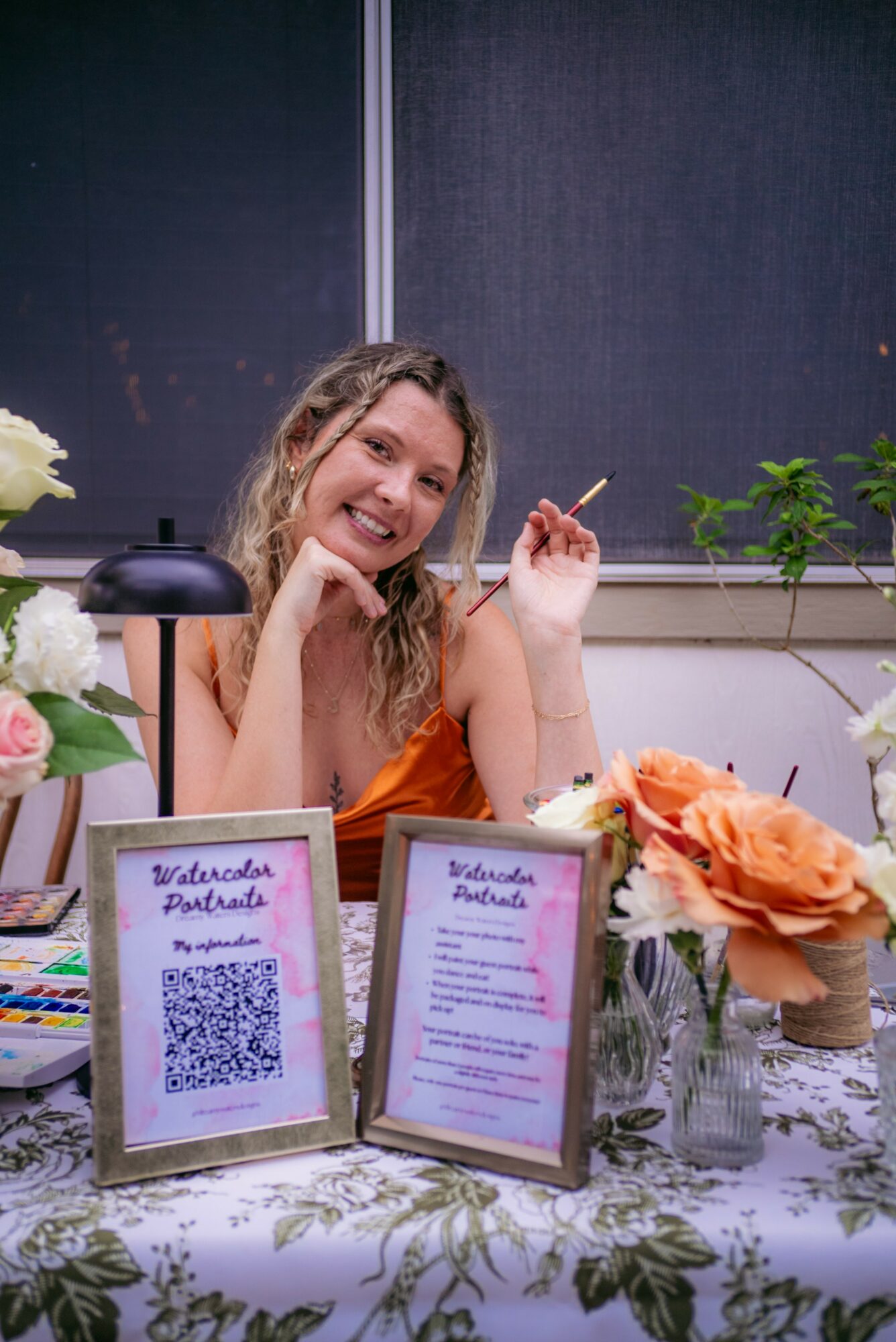 Smiling woman sitting at a table with flowers and framed signs, holding a cigarette, in an indoor setting.