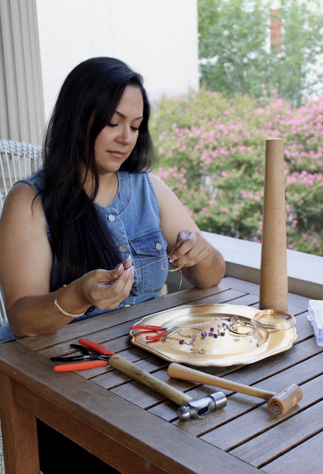Woman with long dark hair in a sleeveless denim top sits at a wooden table, working on a craft project outdoors with pink flowers and trees in background.