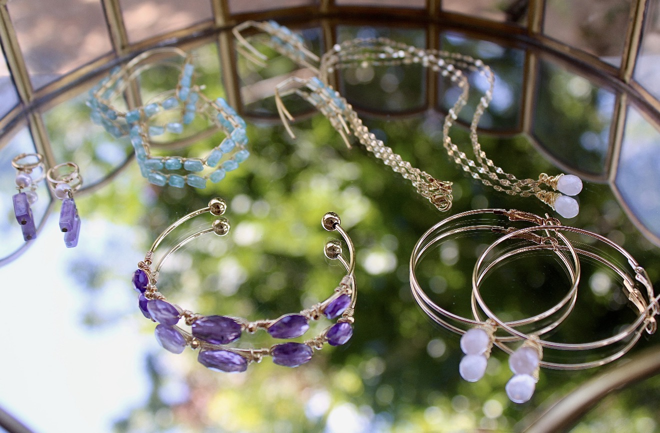 Jewelry including necklaces, bracelets, and earrings displayed on a reflective surface with a blurred outdoor background.