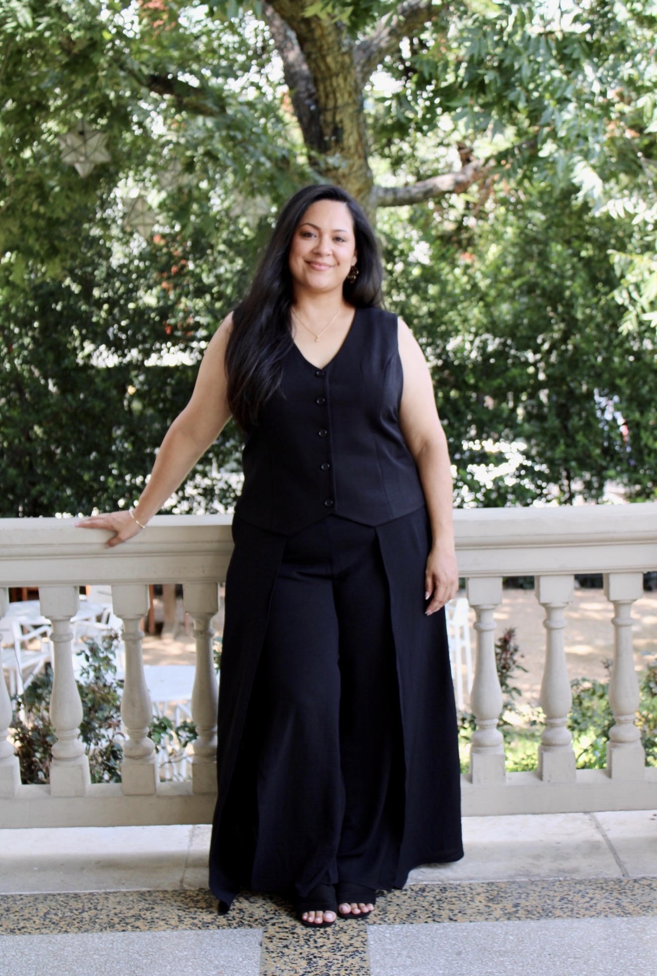 Woman standing outdoors on a balcony with trees in the background, smiling, wearing a black sleeveless top and wide-leg pants.