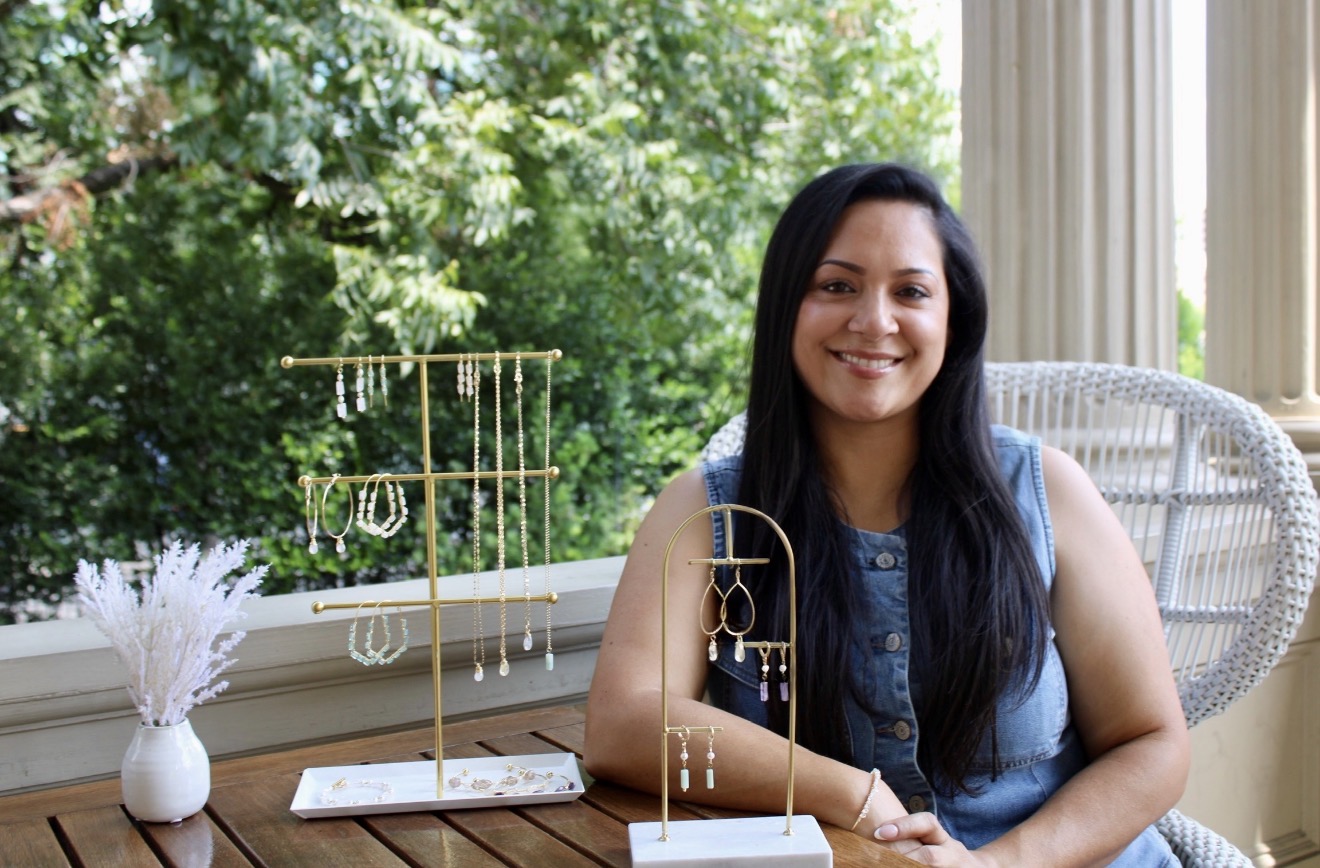 Smiling woman with long dark hair sitting at a table outdoors, with jewelry display and greenery in background.