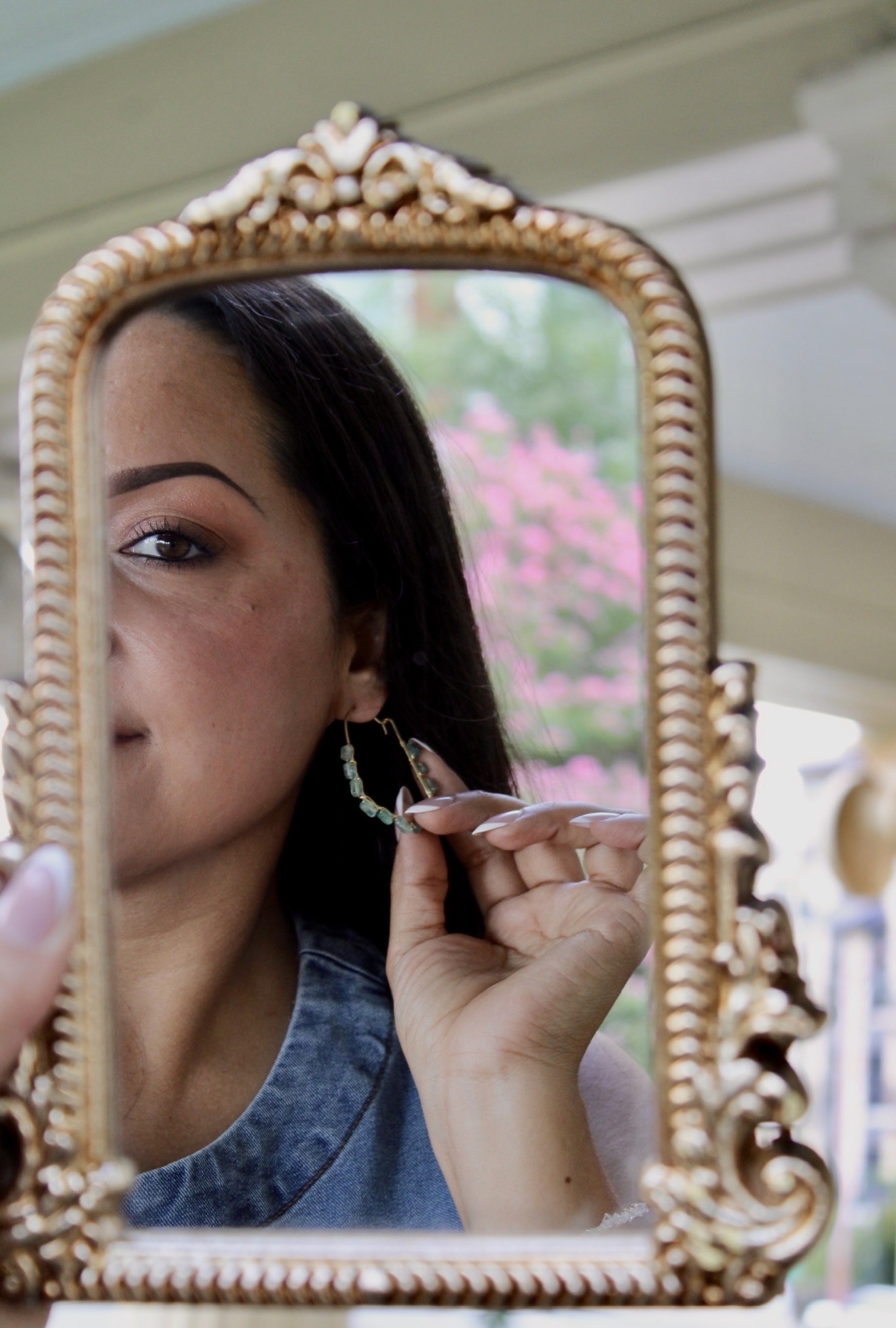 Person looking into ornate mirror, adjusting earring, with pink flowers and greenery in background.