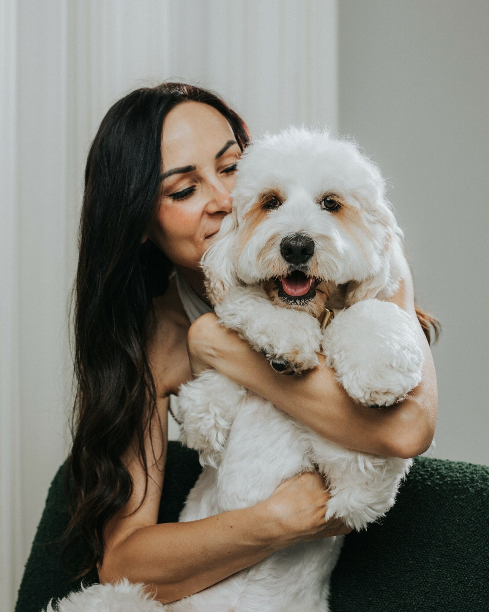 Woman holding a fluffy white dog, smiling, indoors with light-colored curtains in background.