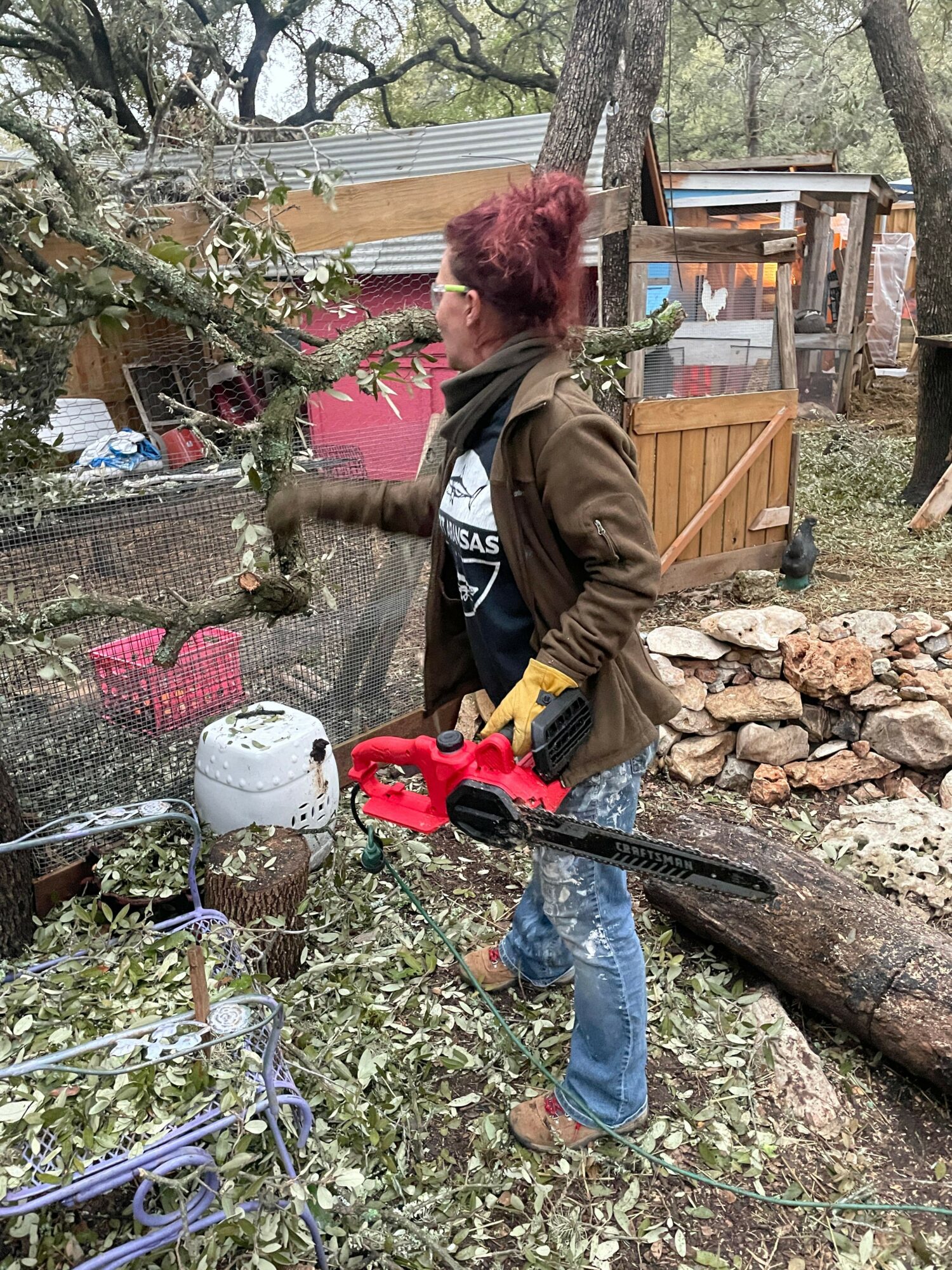 Person with red hair using a chainsaw to cut a log outdoors, surrounded by trees and garden items.