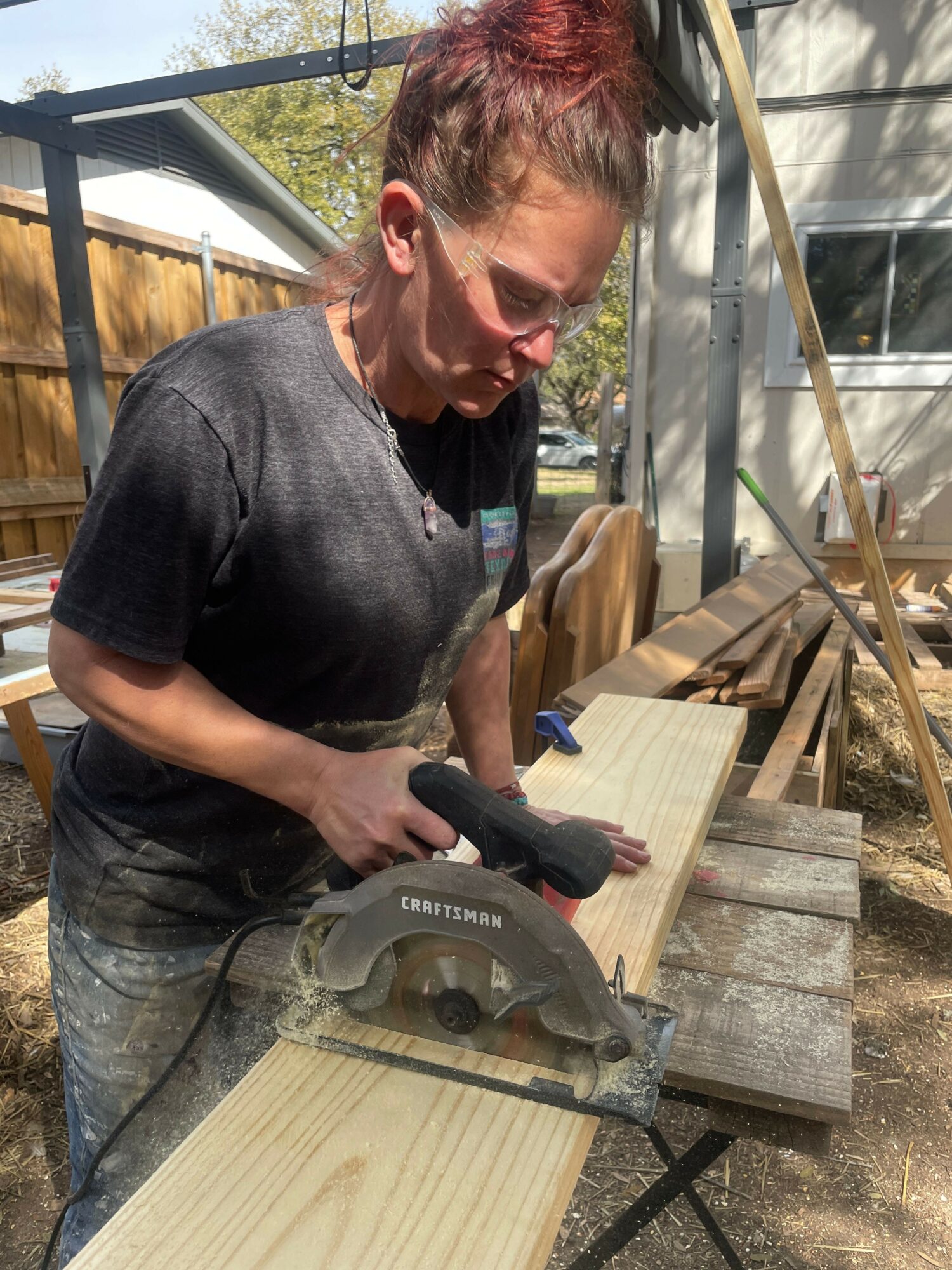 Person with short red hair using a saw on a wooden plank outdoors, surrounded by construction materials and wooden beams.