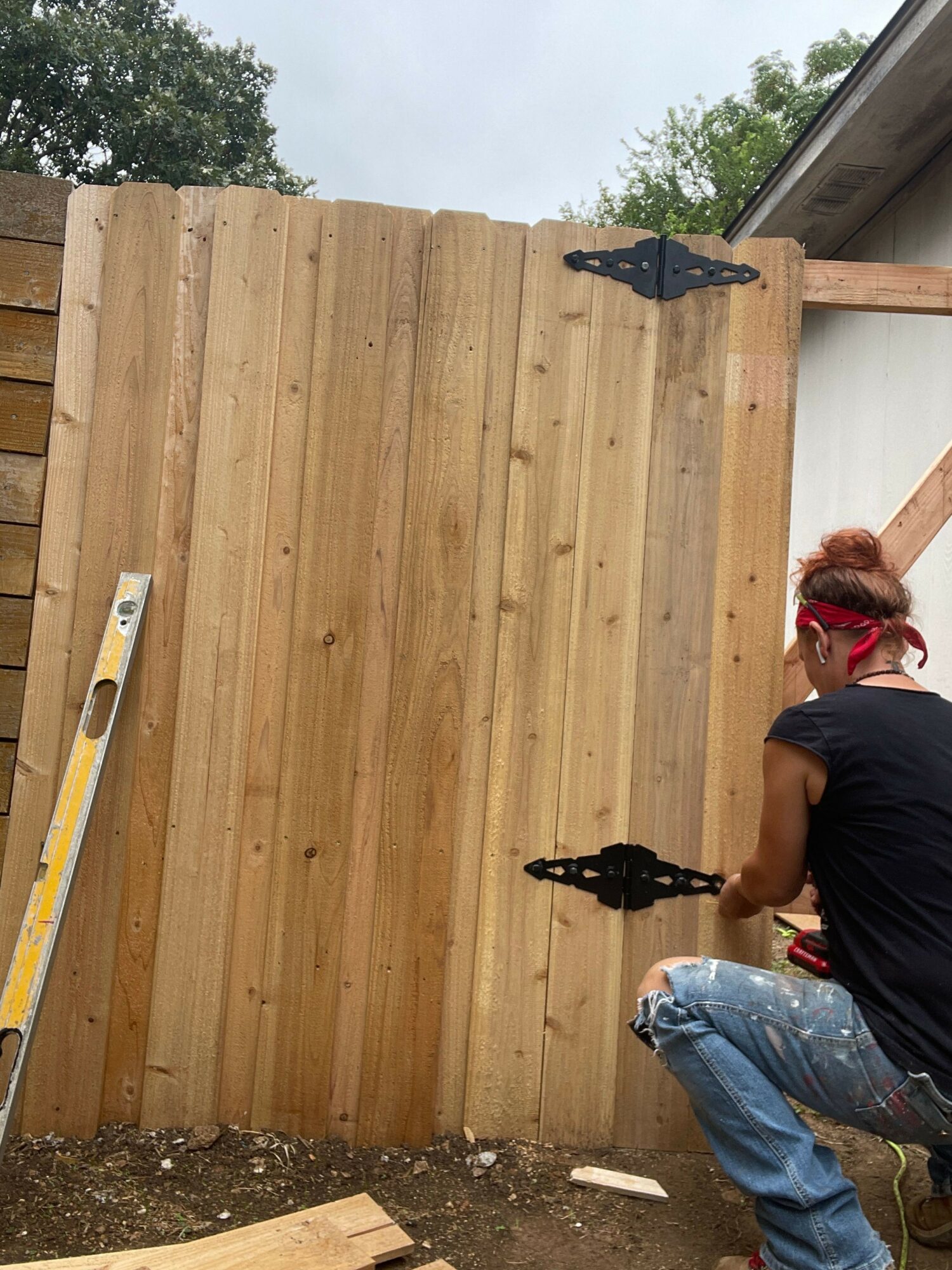 Person working on a wooden fence outdoors, wearing a black shirt and jeans, with trees in the background.