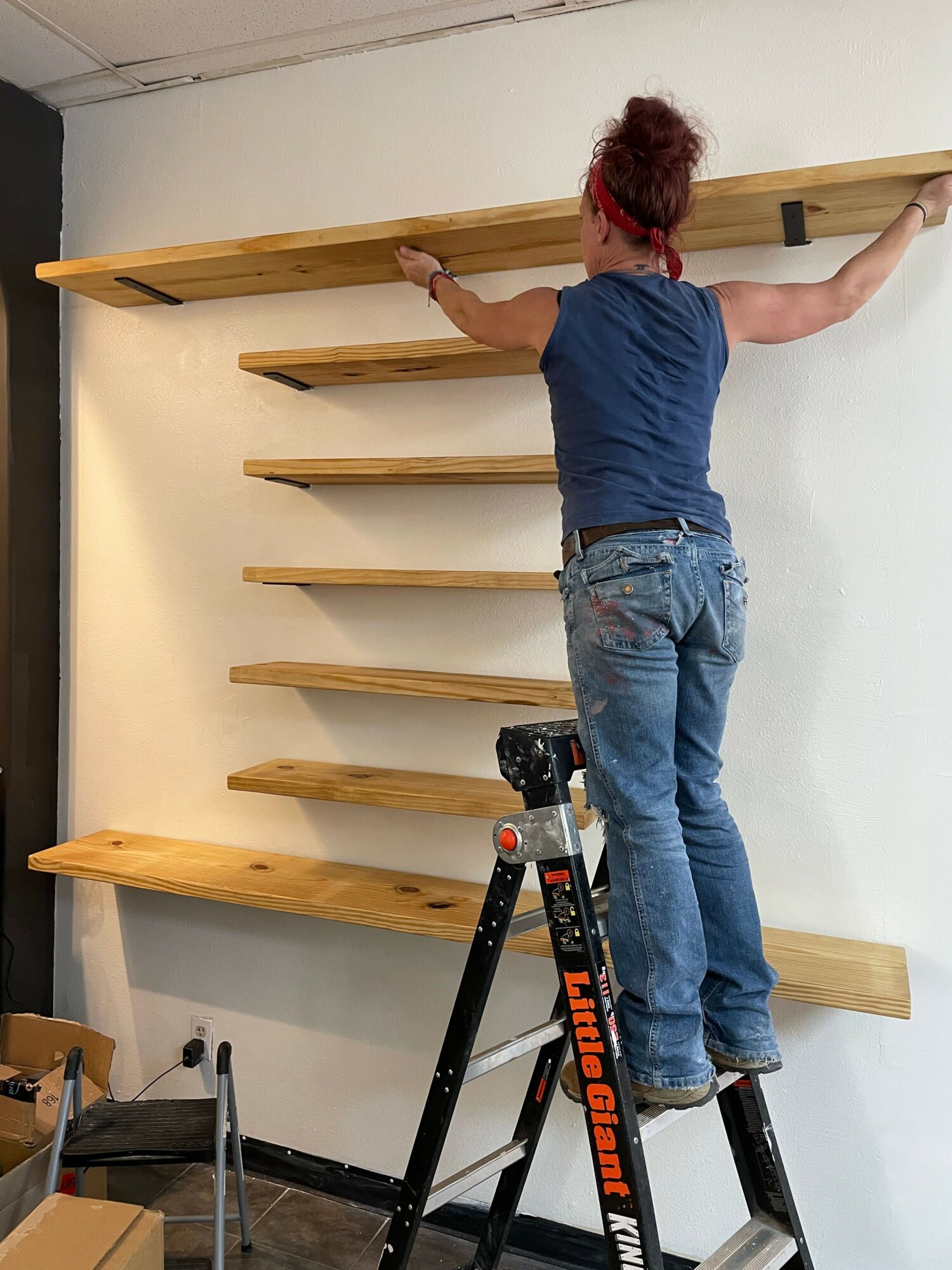 Person lying horizontally on a black Little Giant ladder, leaning against wooden slats on a wall, with head and shoulders supported by the ladder.