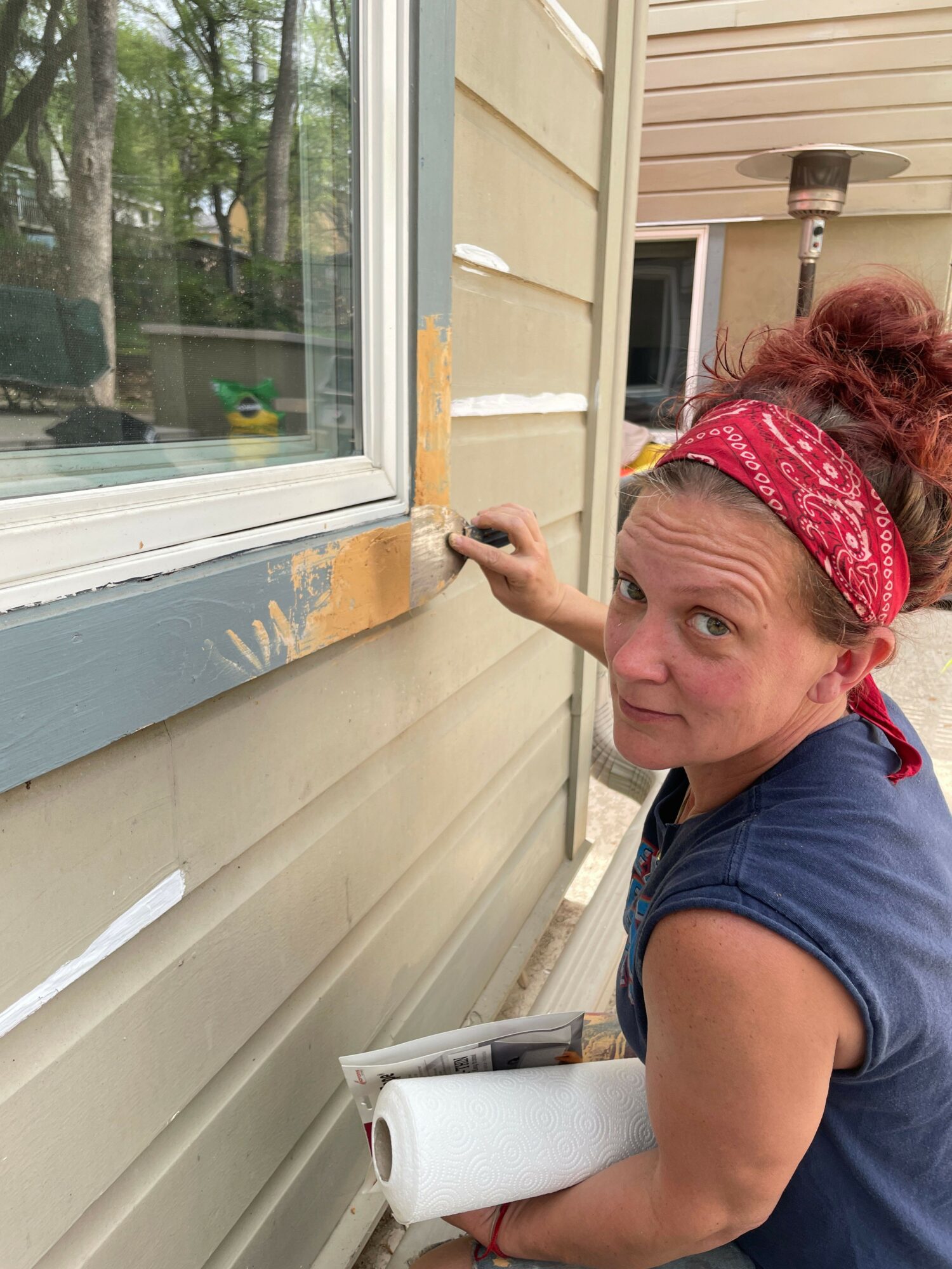 Woman with red bandana cleaning window frame outside, holding a roll of paper, leaning forward, with a house and trees in reflection.