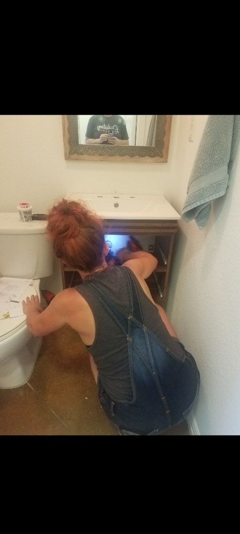 Person with red hair leaning over a toilet, using a laptop on a small white desk, mirror above, towel on the wall.