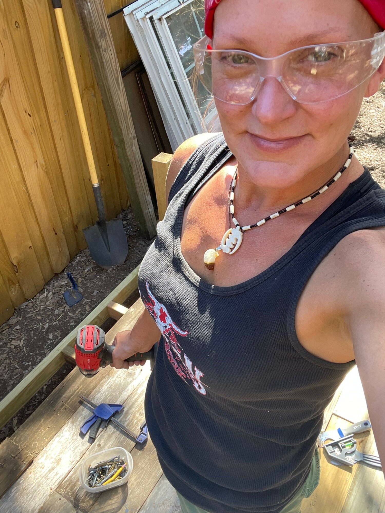 Person with glasses and a necklace, wearing a black shirt, standing outdoors near a wooden structure, with tools and a soda can nearby.