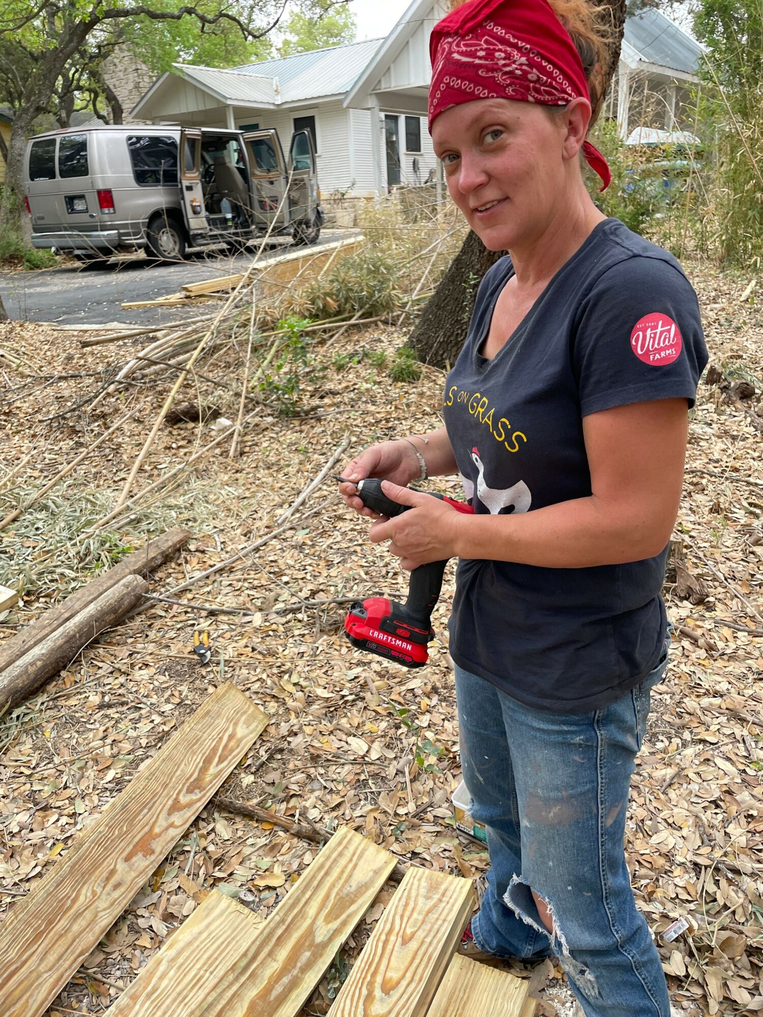 Woman with a red bandana, dark shirt, and jeans working outdoors on a wooden project, holding a tool, with a vehicle and trees in background.