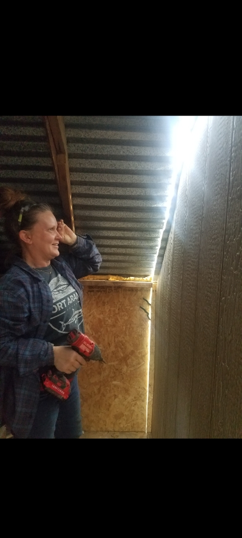 Woman smiling, holding a red object, standing in a wooden and metal structure, with light coming from above.