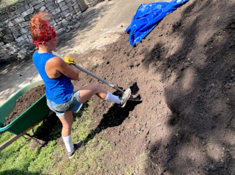 Child with curly red hair in a blue tank top and shorts shoveling dirt outdoors, with a wheelbarrow nearby.