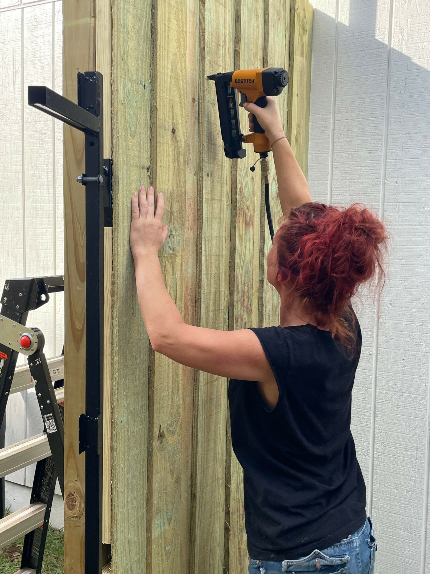 Person with red hair using a power tool on wooden planks outdoors, lying on their stomach, wearing a black shirt and jeans.