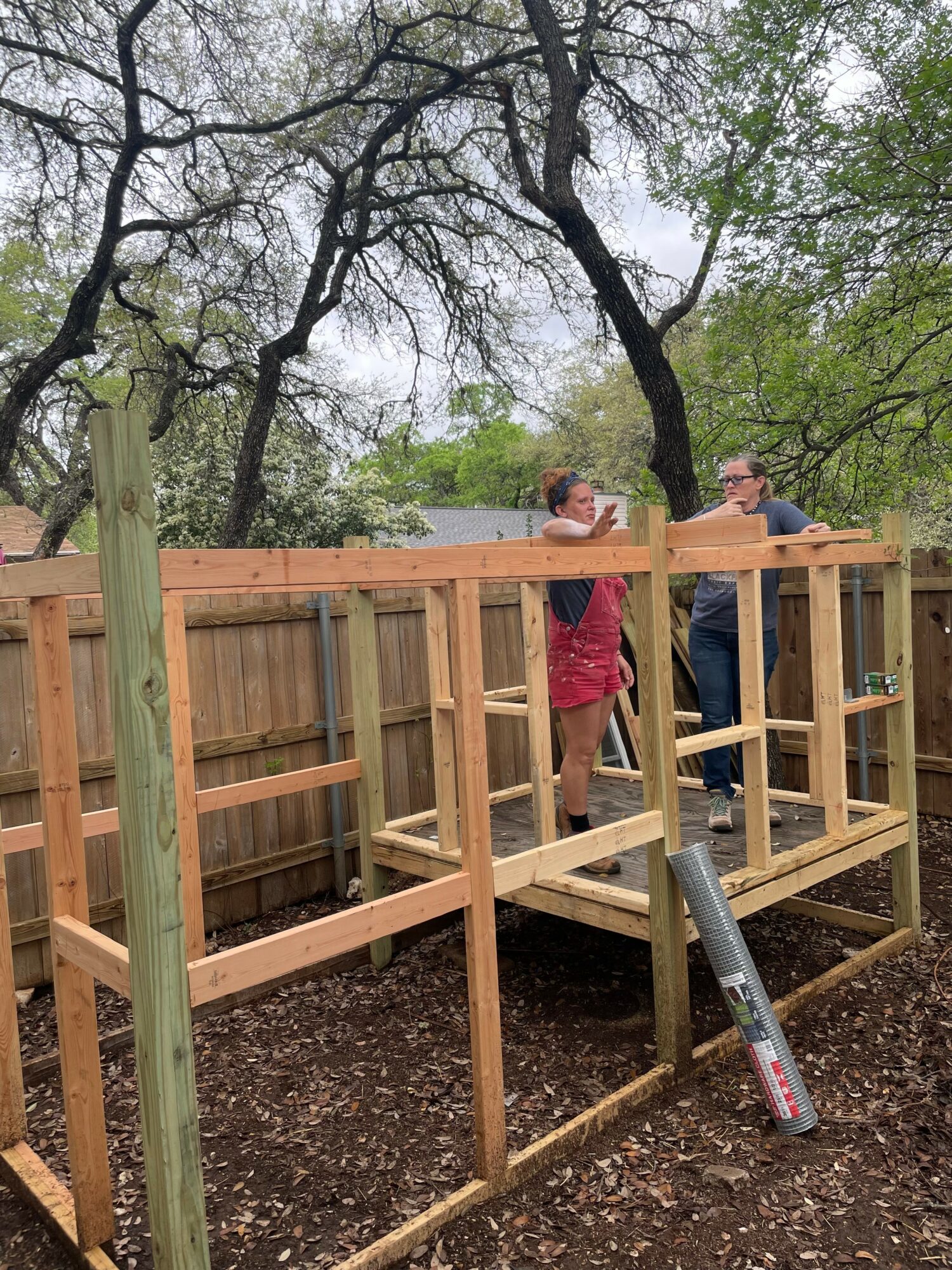 Two people working on a wooden deck in a backyard with trees and a fence, one person in a red dress, the other in a gray shirt.