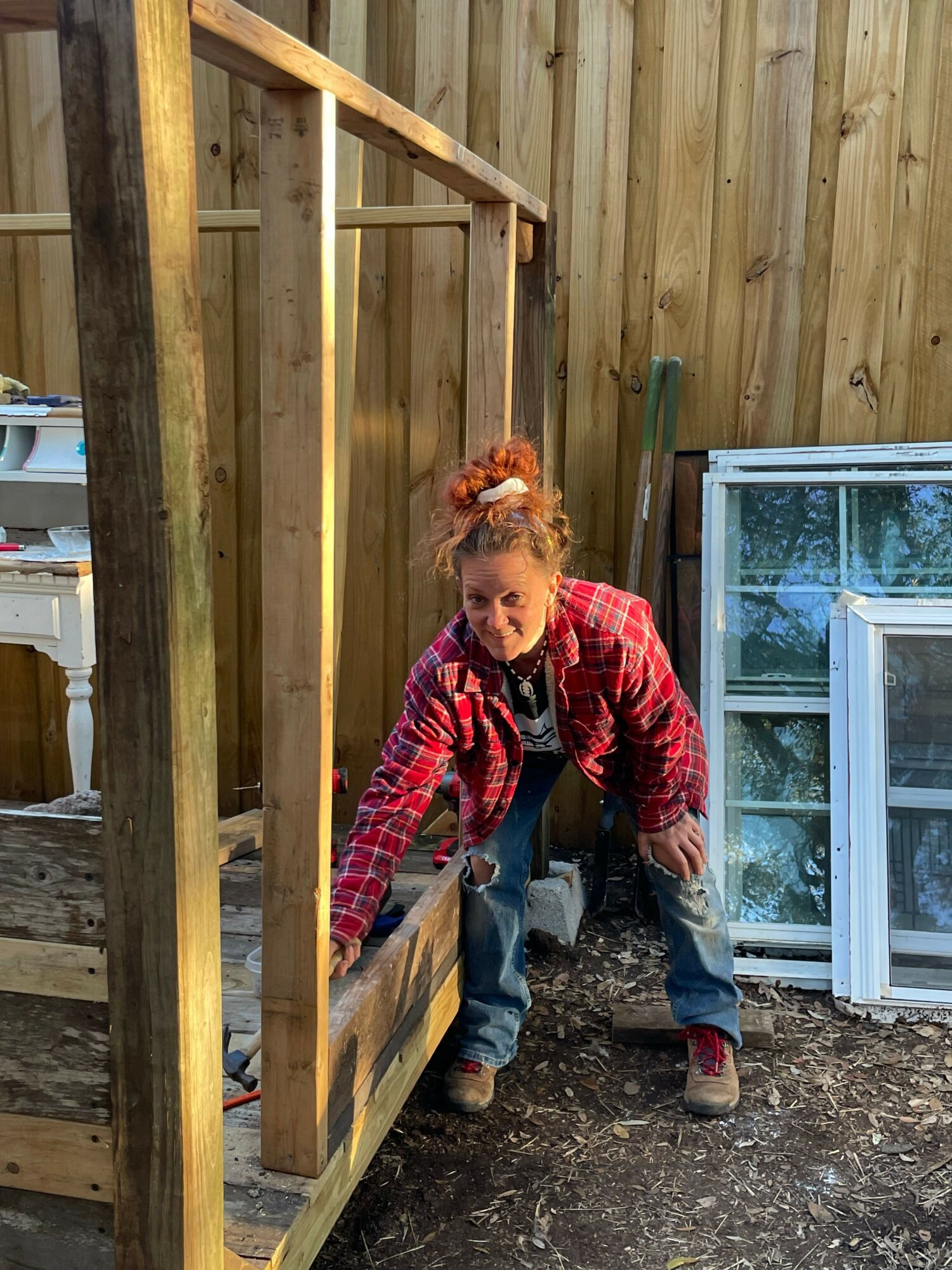 Child with red hair and red jacket crouches next to a wooden structure outdoors, with a window and wooden wall in background.