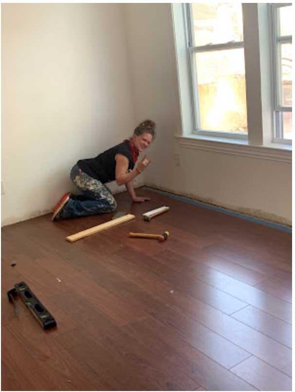 Person kneeling on floor installing wooden flooring near window, tools and measuring tape nearby, bright room with sunlight.