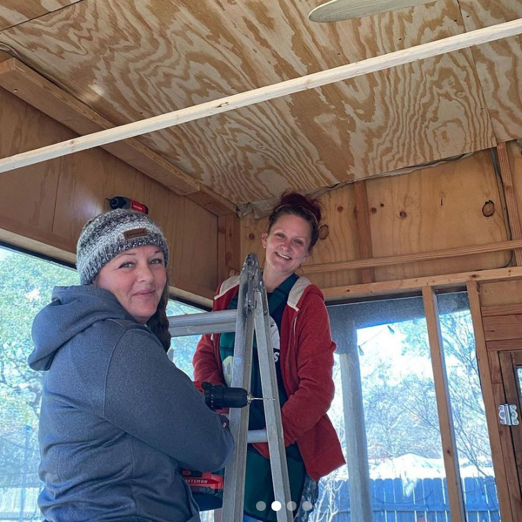 Two women inside a wooden cabin, smiling, with snow visible outside through windows.