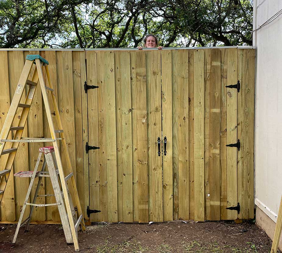 Person peeking over a newly installed wooden fence with a ladder nearby, outdoors with trees in the background.