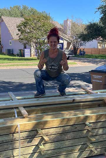Person crouching outdoors on wooden planks, smiling, with trees and houses in the background.