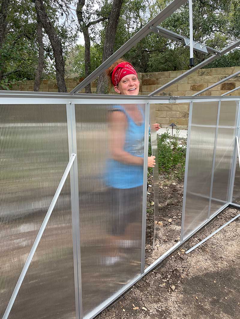 Person with red headband smiling inside a transparent greenhouse structure, outdoors with trees and a brick wall in background.