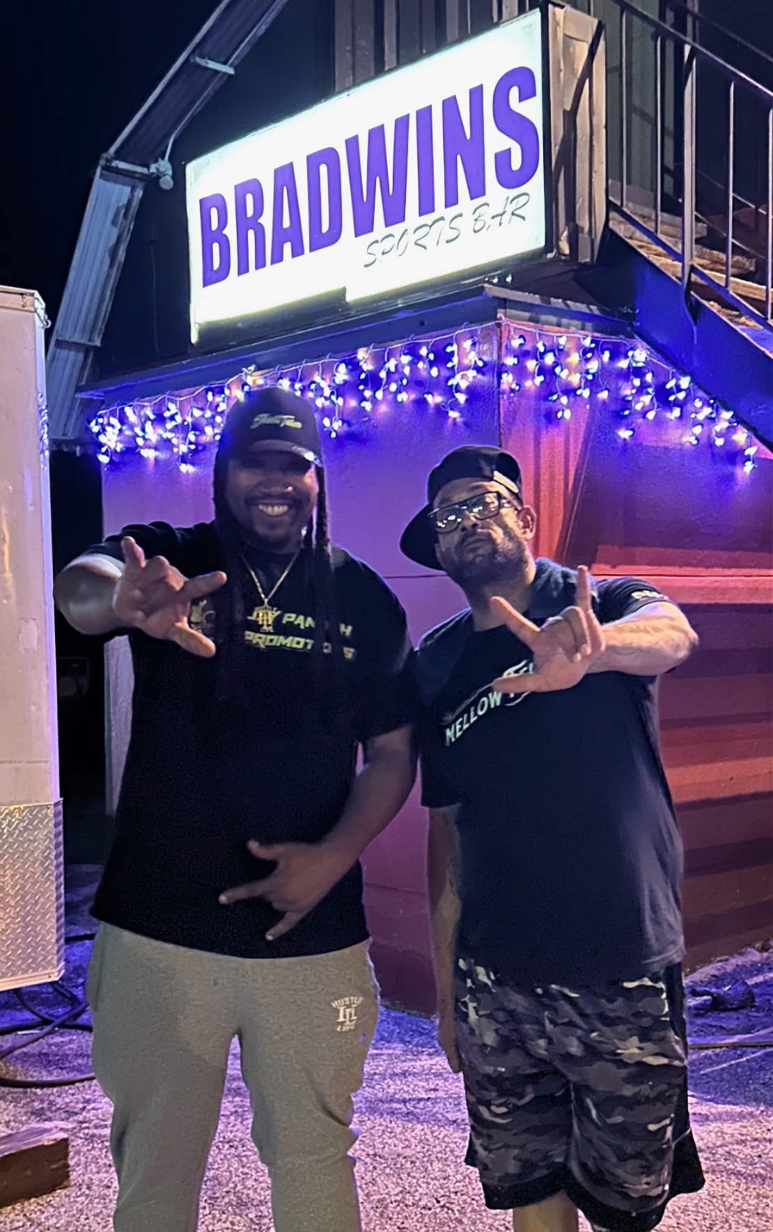 Two men standing in front of a sign that reads 'BRADWINS SPORTS BAR,' smiling and making hand gestures, with purple lighting and decorations.