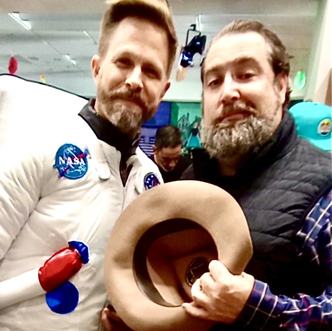 Two men with beards, one in a NASA spacesuit, holding a hat, in an indoor setting with people in the background.