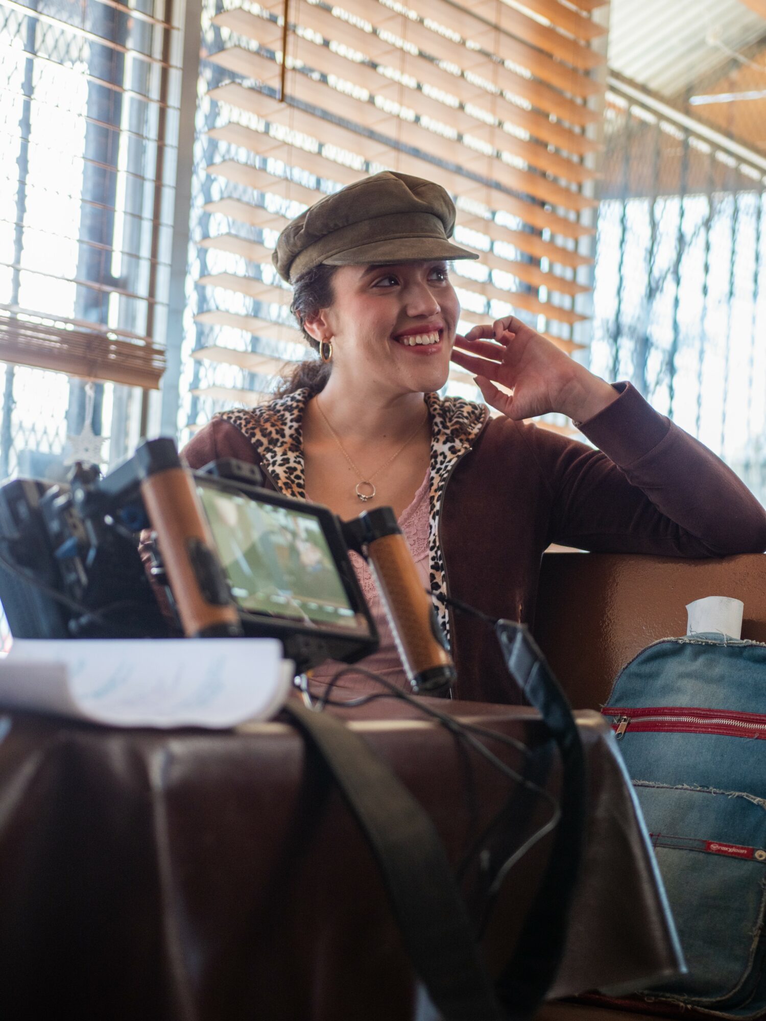 Woman smiling, wearing a hat, sitting indoors near window blinds, with camera equipment and a backpack on table.