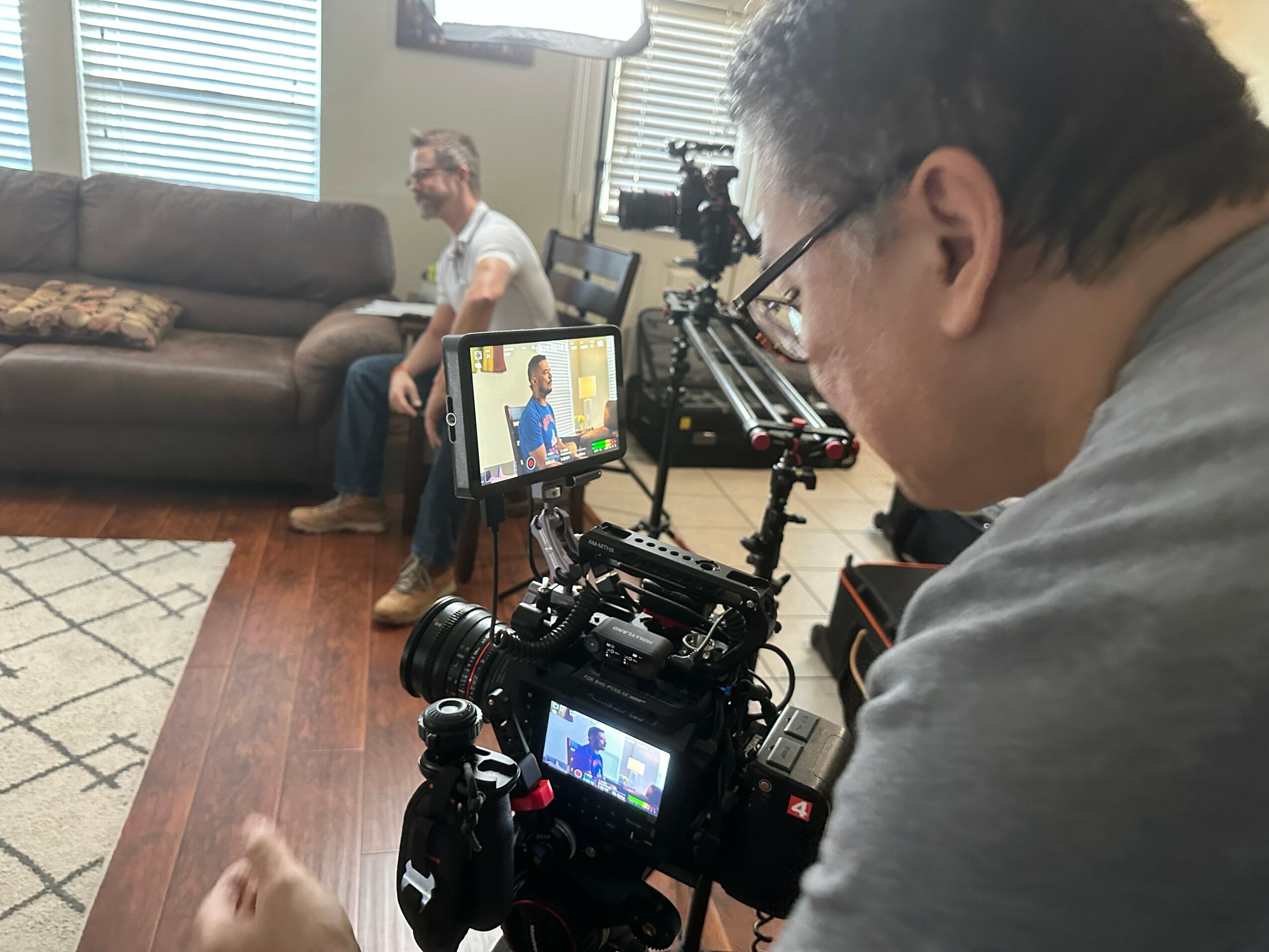 Filmmaker filming a person sitting on a sofa in a room with windows, camera equipment visible, and a monitor showing the scene.