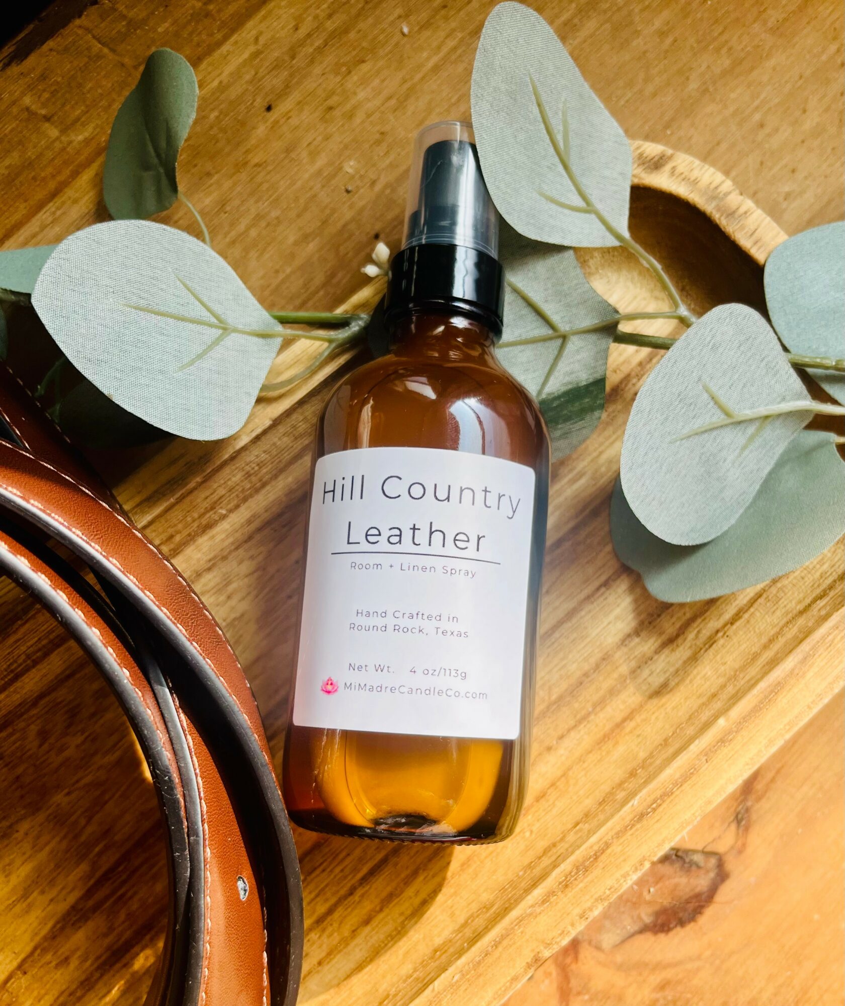 Amber spray bottle labeled 'Hill Country Leather' on wooden surface with green leaves around it.