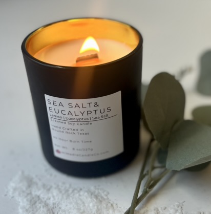 Lit candle in black jar with label, next to eucalyptus leaves on white surface.