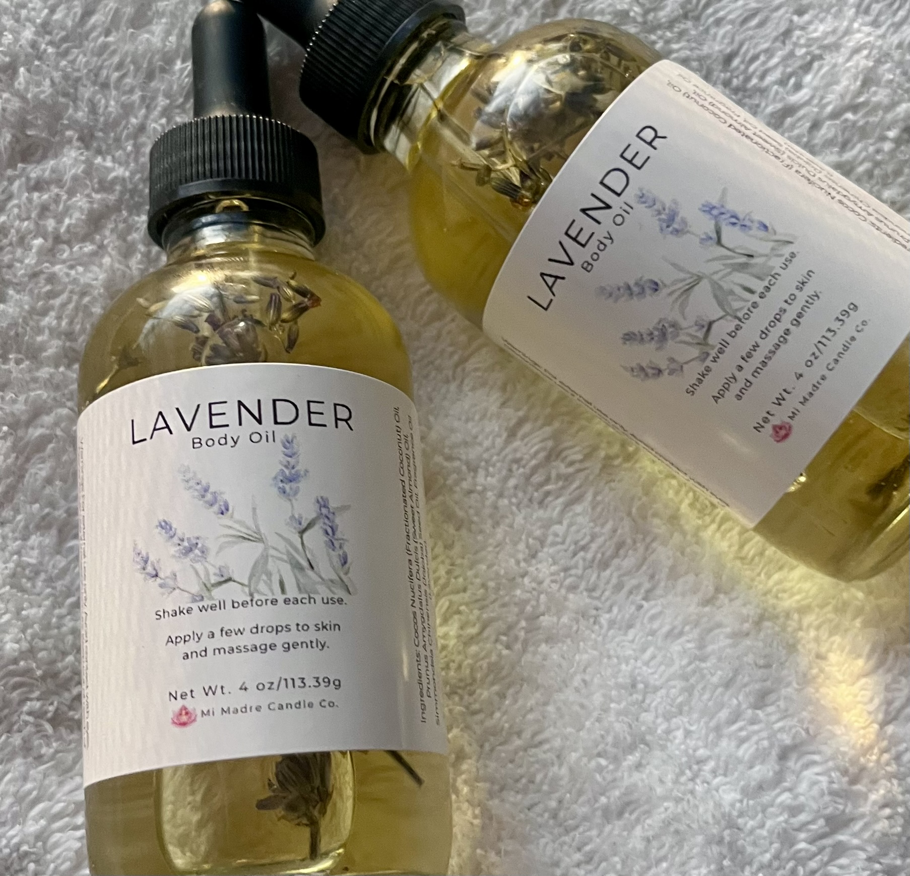 Two bottles of lavender body oil with black caps on a soft gray surface, labels showing lavender illustrations and text.
