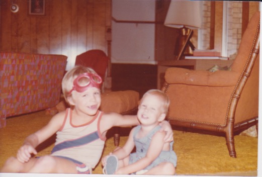 Two children sitting on the floor in a living room, one wearing goggles and the other smiling.