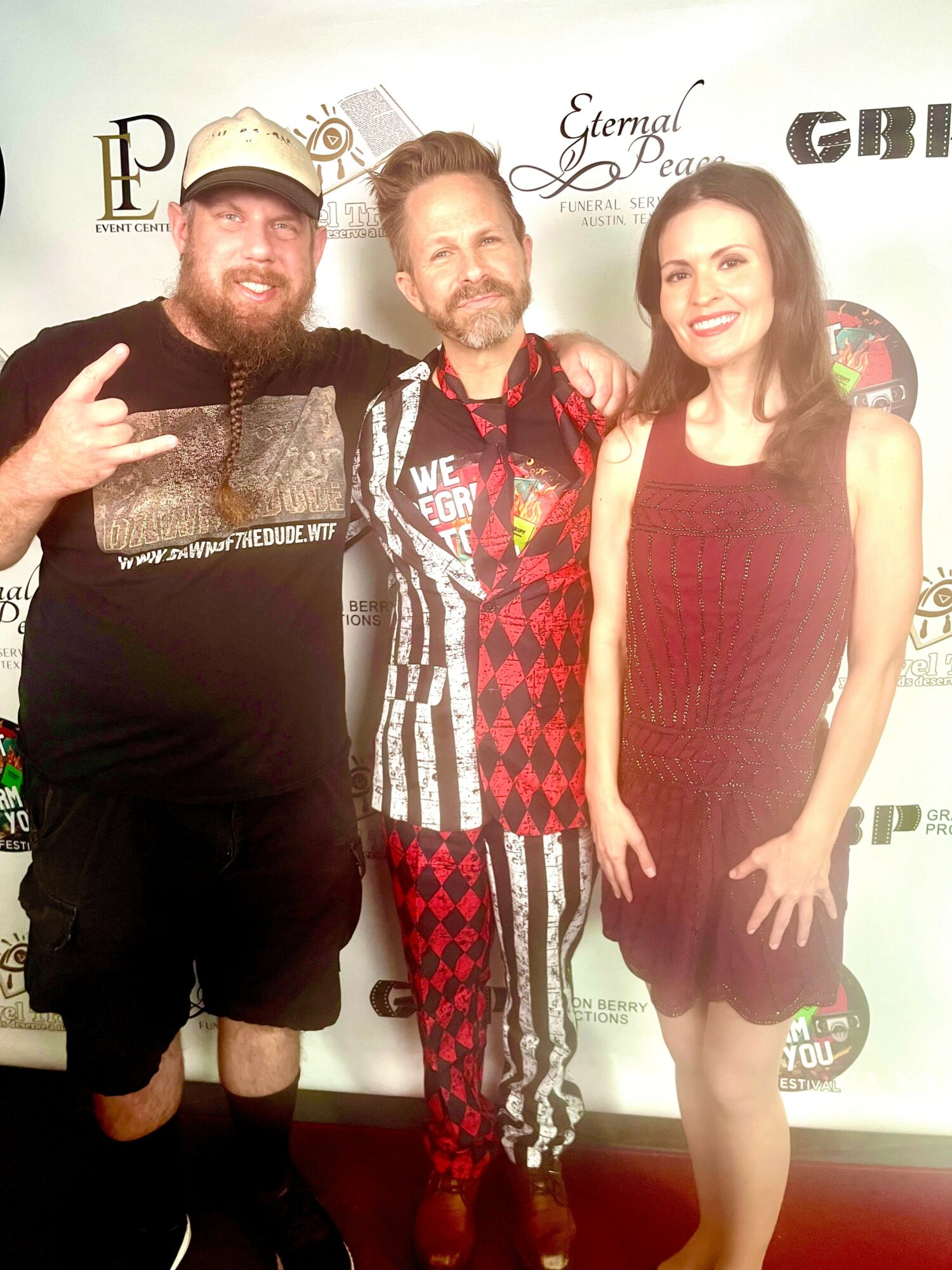 Three people standing together, smiling, in front of a backdrop with logos, one making a peace sign, two women and one man.