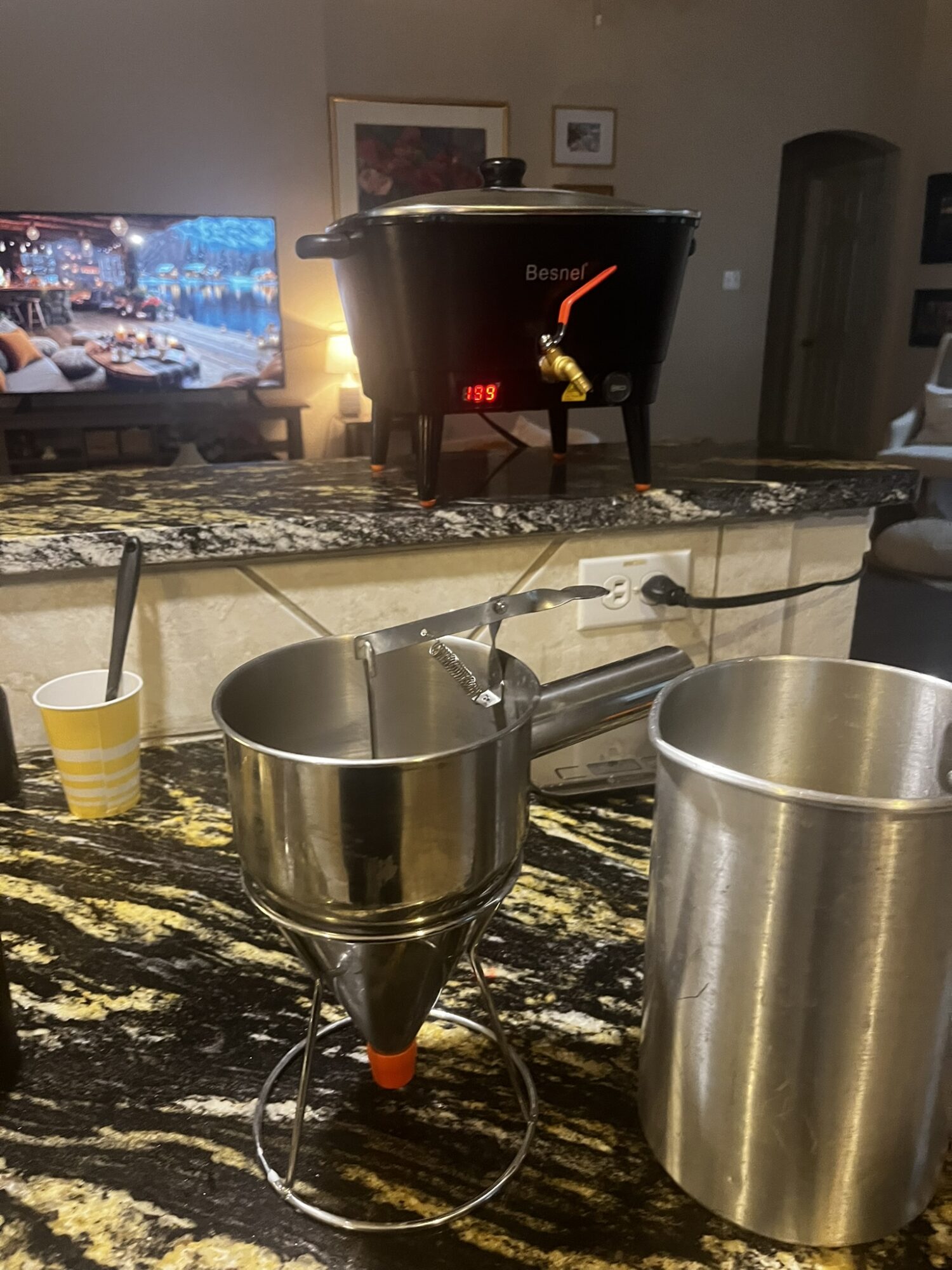 Pouring liquid into a metal funnel on a kitchen counter with a large metal container nearby, in a home setting.