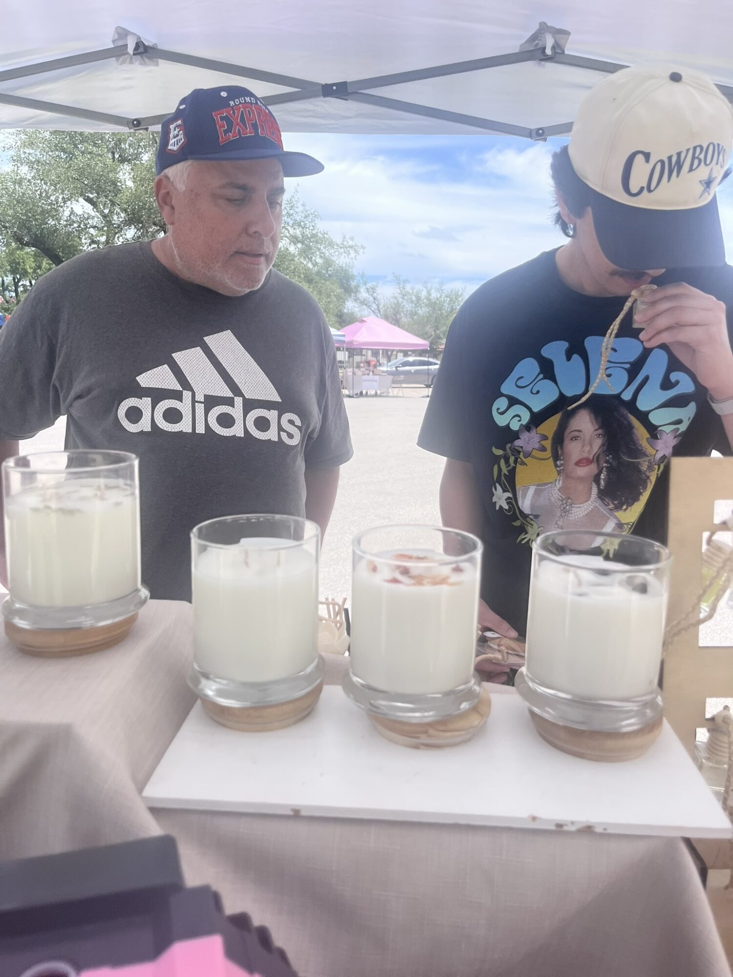 Two men sit at a table with four glasses of milk, outdoors under a canopy, with trees and tents in background.