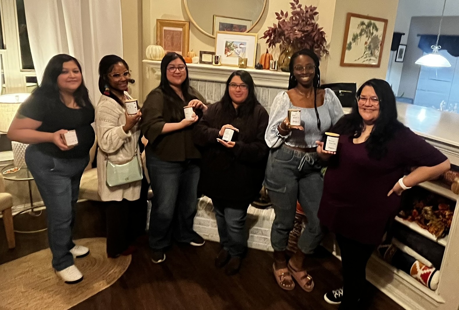 Six women standing indoors, holding small boxes, in a room with framed pictures and a fireplace.