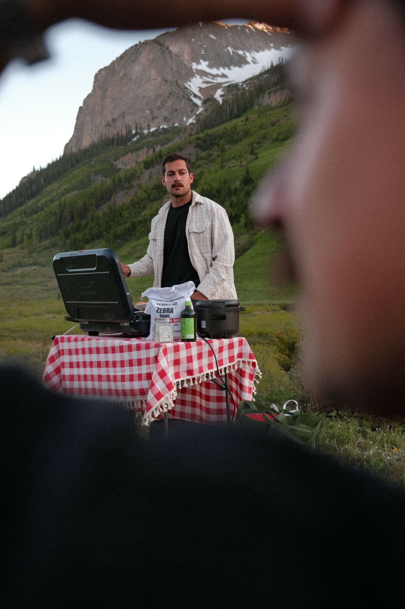 Person standing outdoors near a mountain with snow, with a table and a person taking a photo or video.