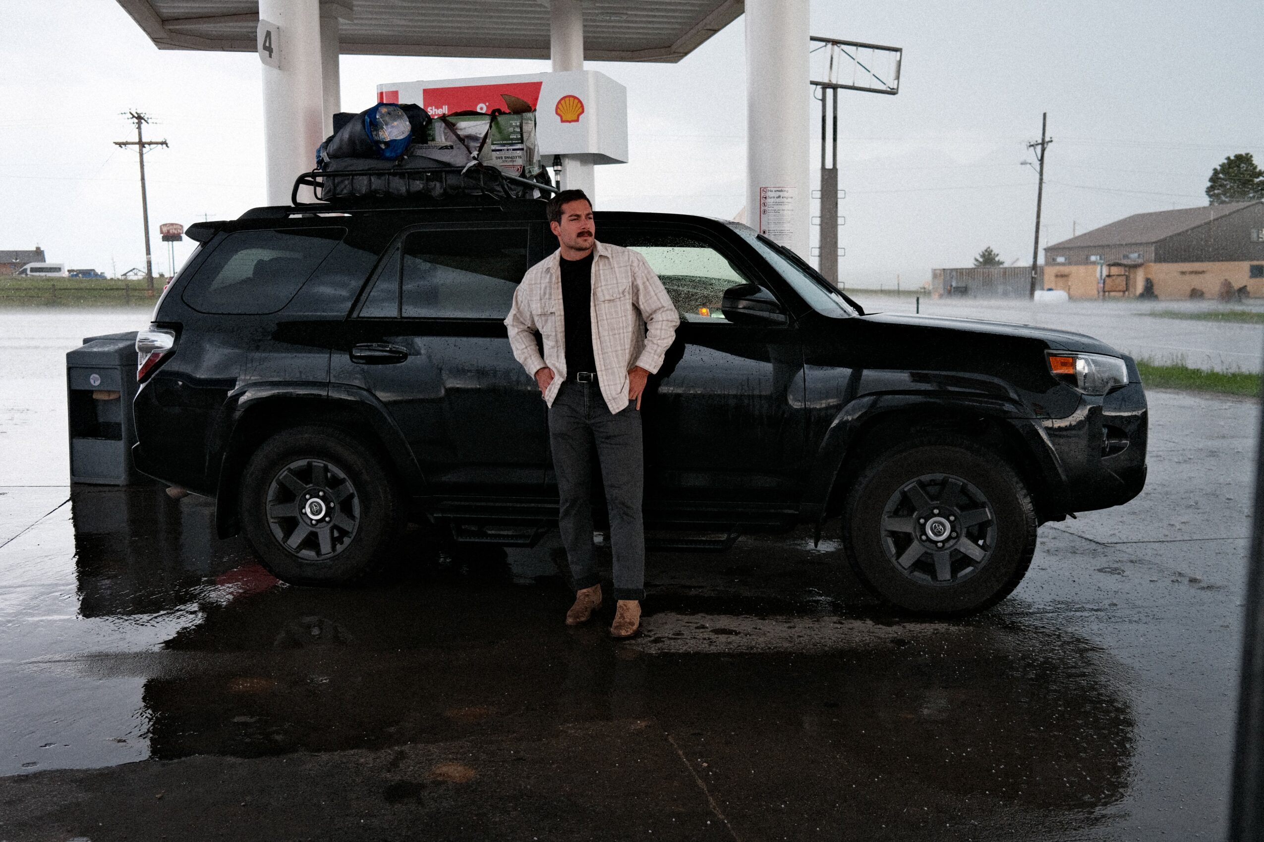 Man standing next to a black SUV at a gas station on a rainy day, with a luggage rack on top.