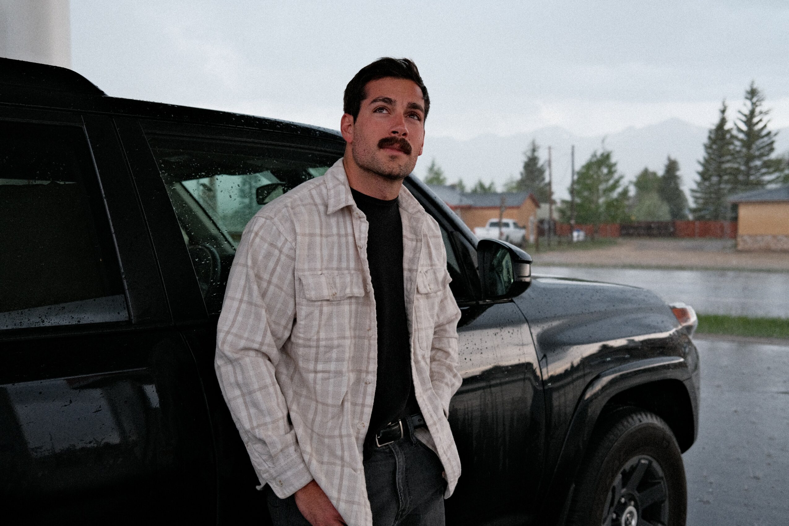 Man with dark hair and mustache stands next to a black vehicle on a rainy day, with trees and buildings in the background.