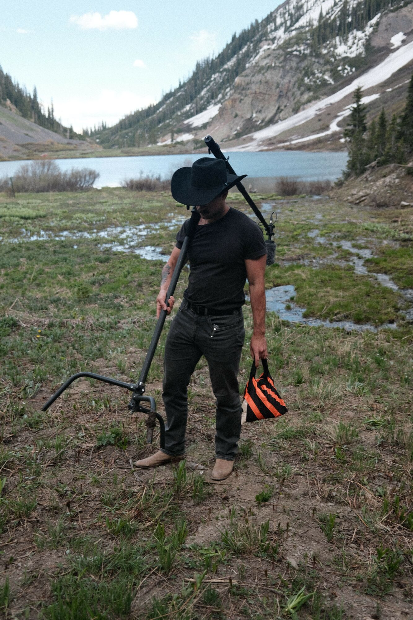 Person standing outdoors near water, holding a rifle, wearing a black hat and dark clothing, with a landscape of trees and snow in the background.