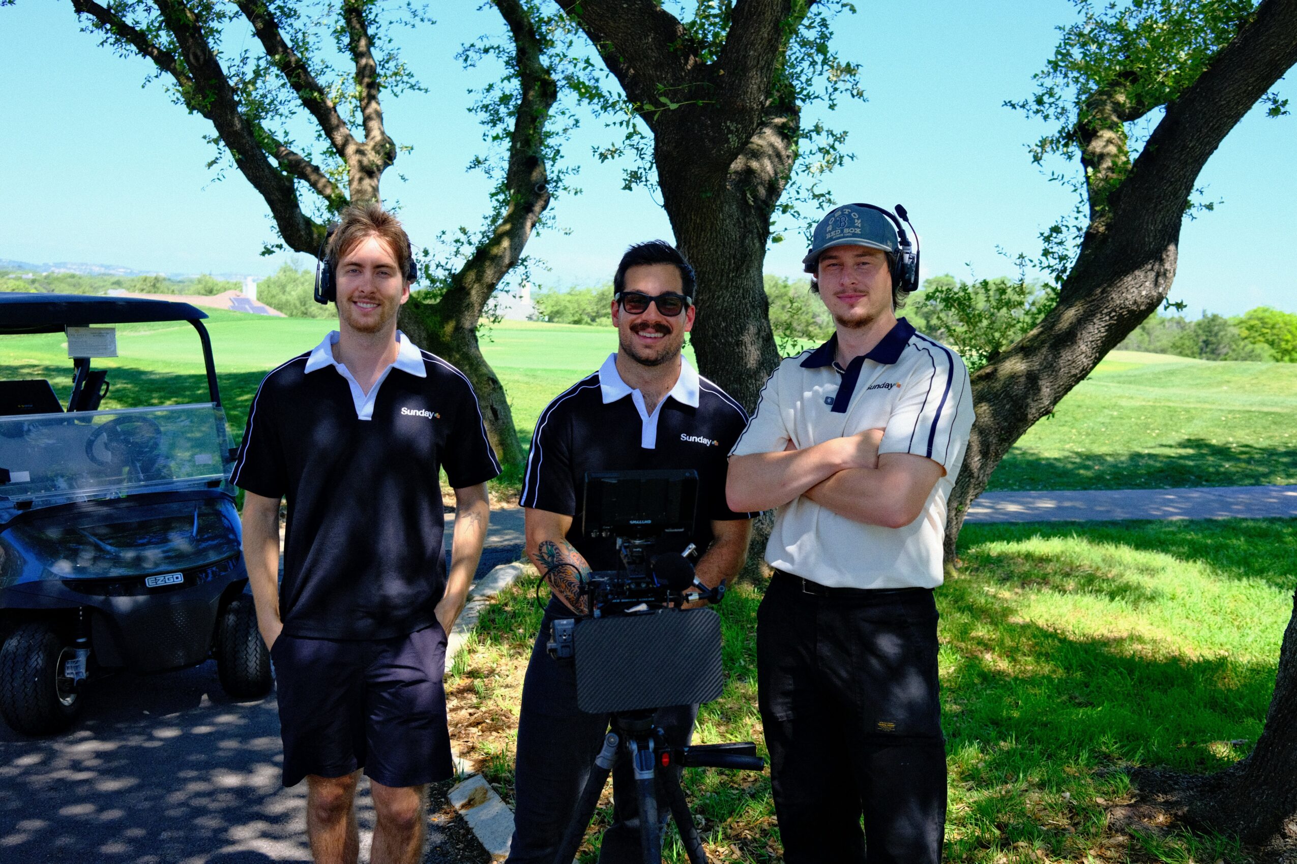 Three men standing outdoors under a tree, smiling, with a golf cart in the background, on a sunny day.