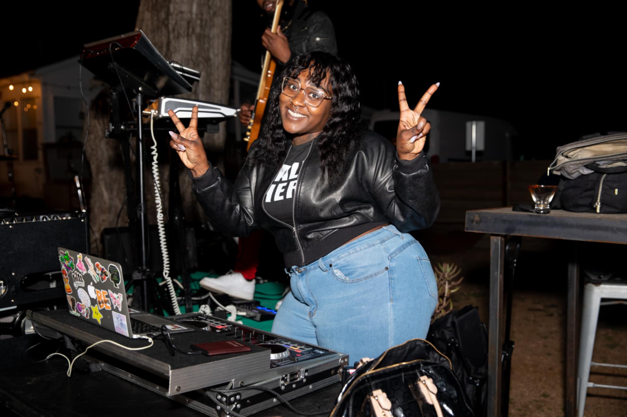 Woman with curly hair smiling and making a peace sign, standing behind DJ equipment outdoors at night.