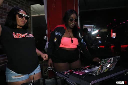 Two women standing near DJ equipment in a dimly lit indoor setting, one wearing sunglasses and a black shirt, the other in a pink and black top.