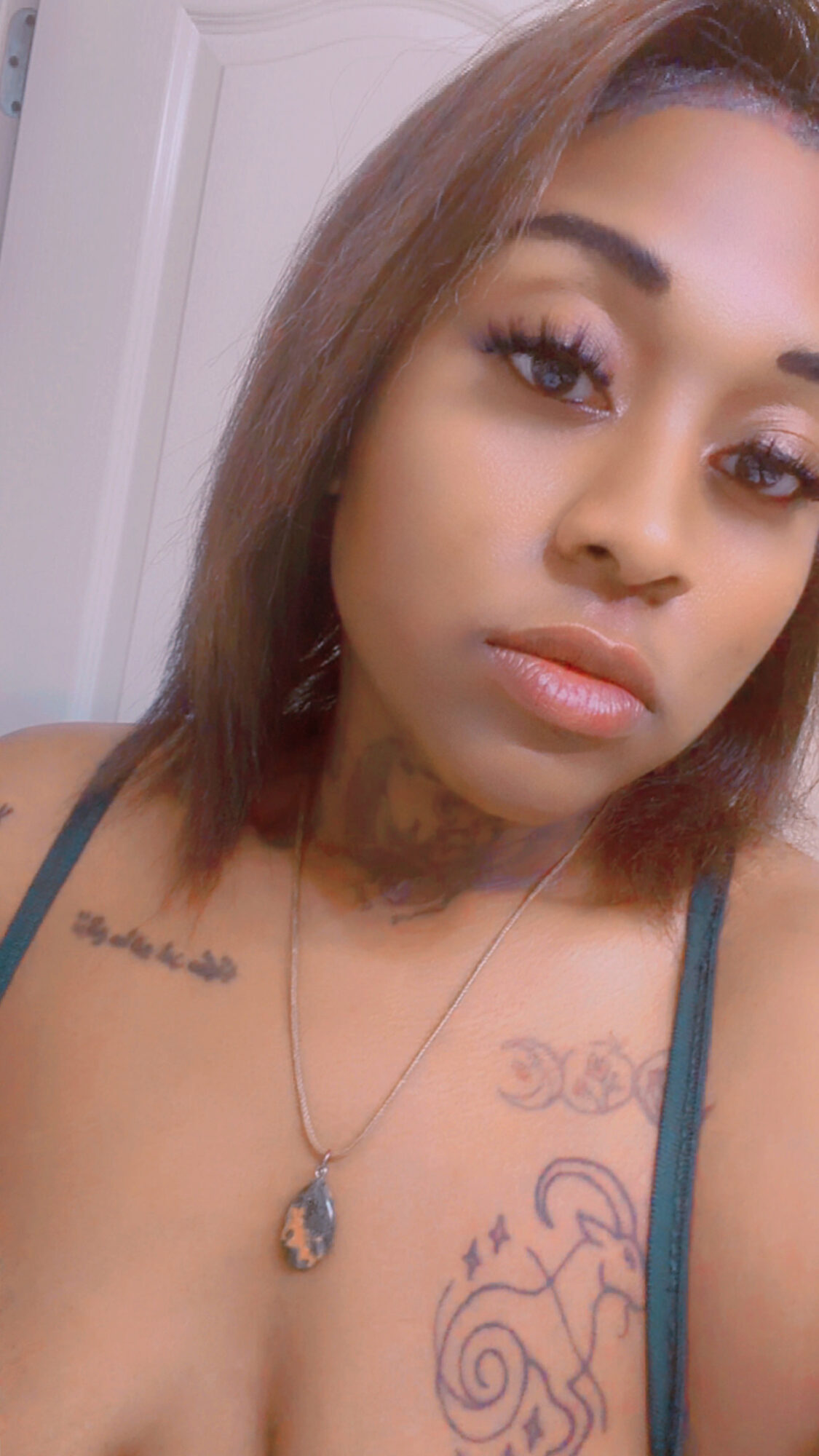Close-up of a woman with shoulder-length hair, tattoos on her chest, wearing a necklace, looking at the camera.