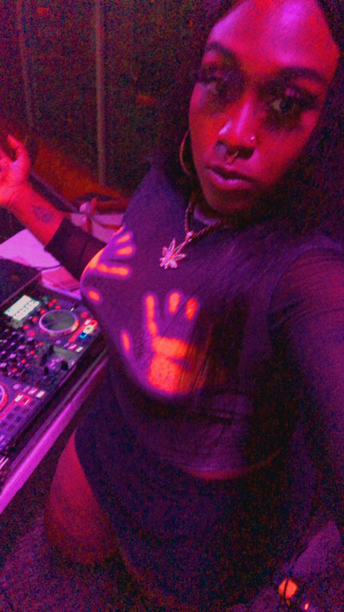 Person with long dark hair in a dark setting, DJ equipment visible, colorful lighting on face and chest.