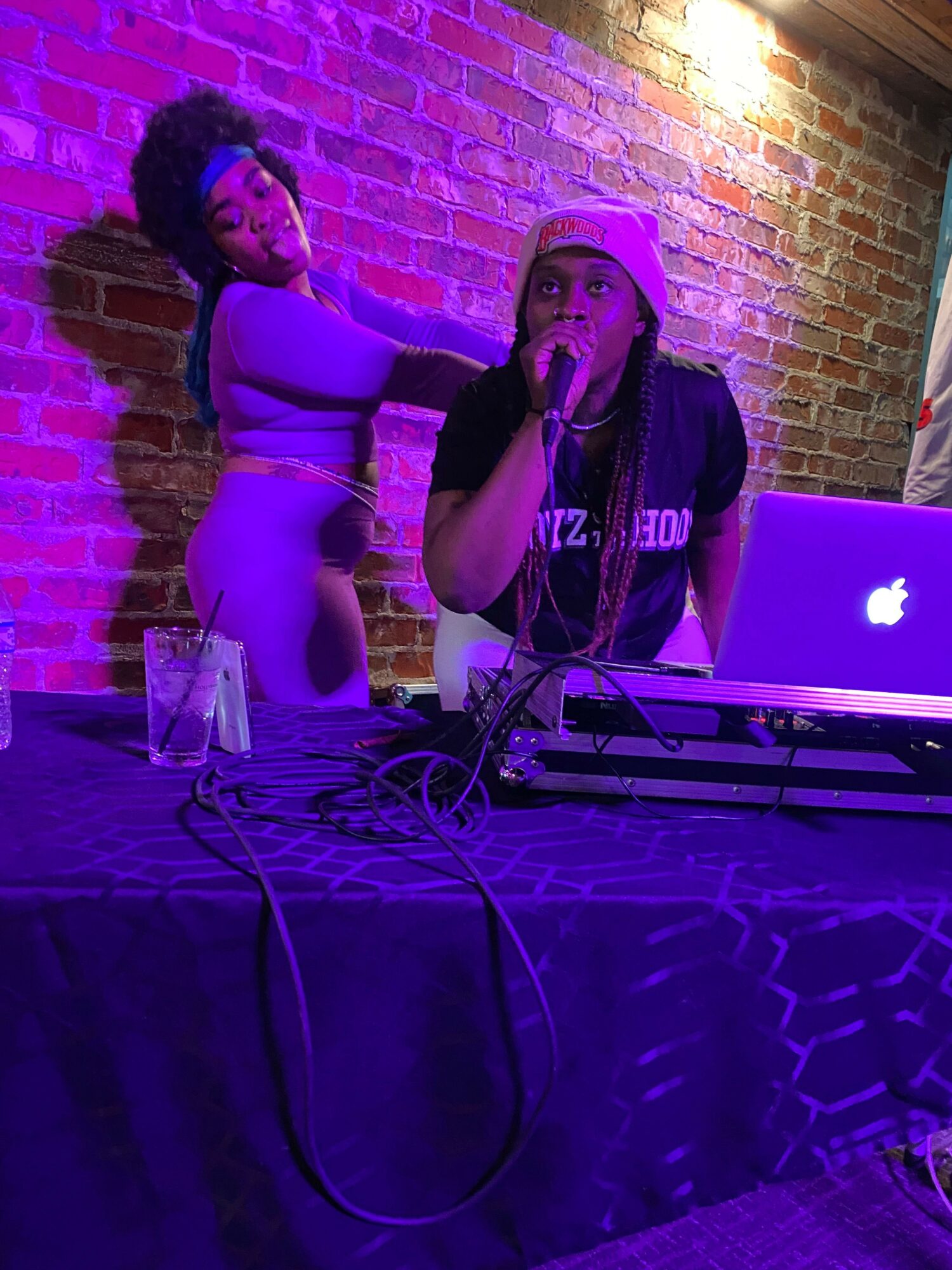 Two women perform on stage with a brick wall background, purple lighting, and DJ equipment nearby.